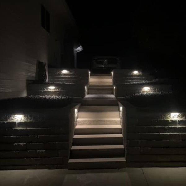 A set of outdoor stone steps illuminated at night by modern integrated wall lights, leading up between tiered retaining walls toward a driveway and a parked vehicle.