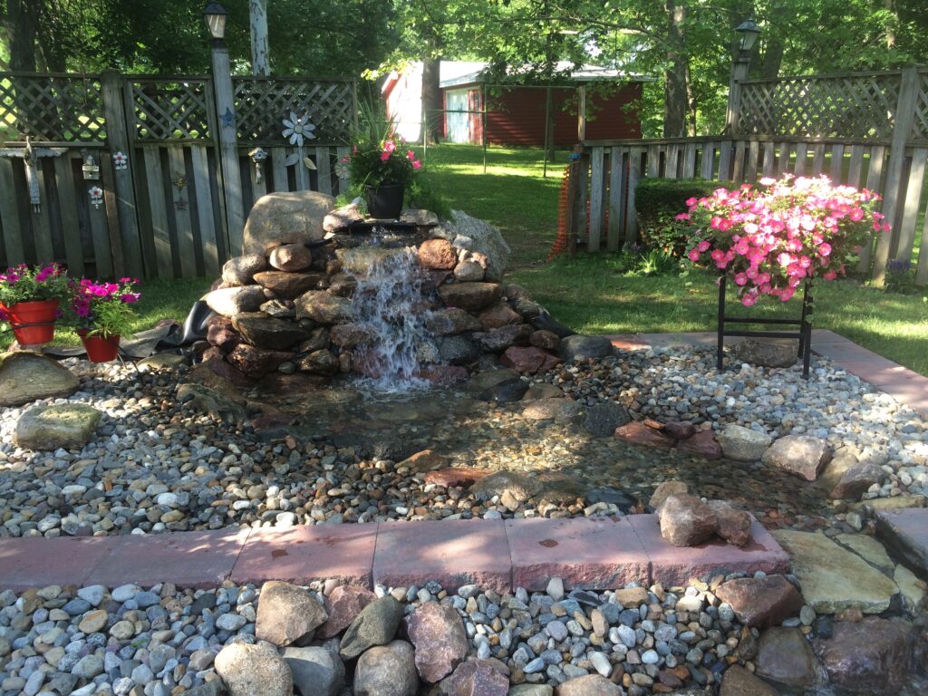 Water feature with cascading waterfall, surrounded by colorful flowers in pots, decorative stones, and a lush green landscape, enhancing a low-maintenance yard design.