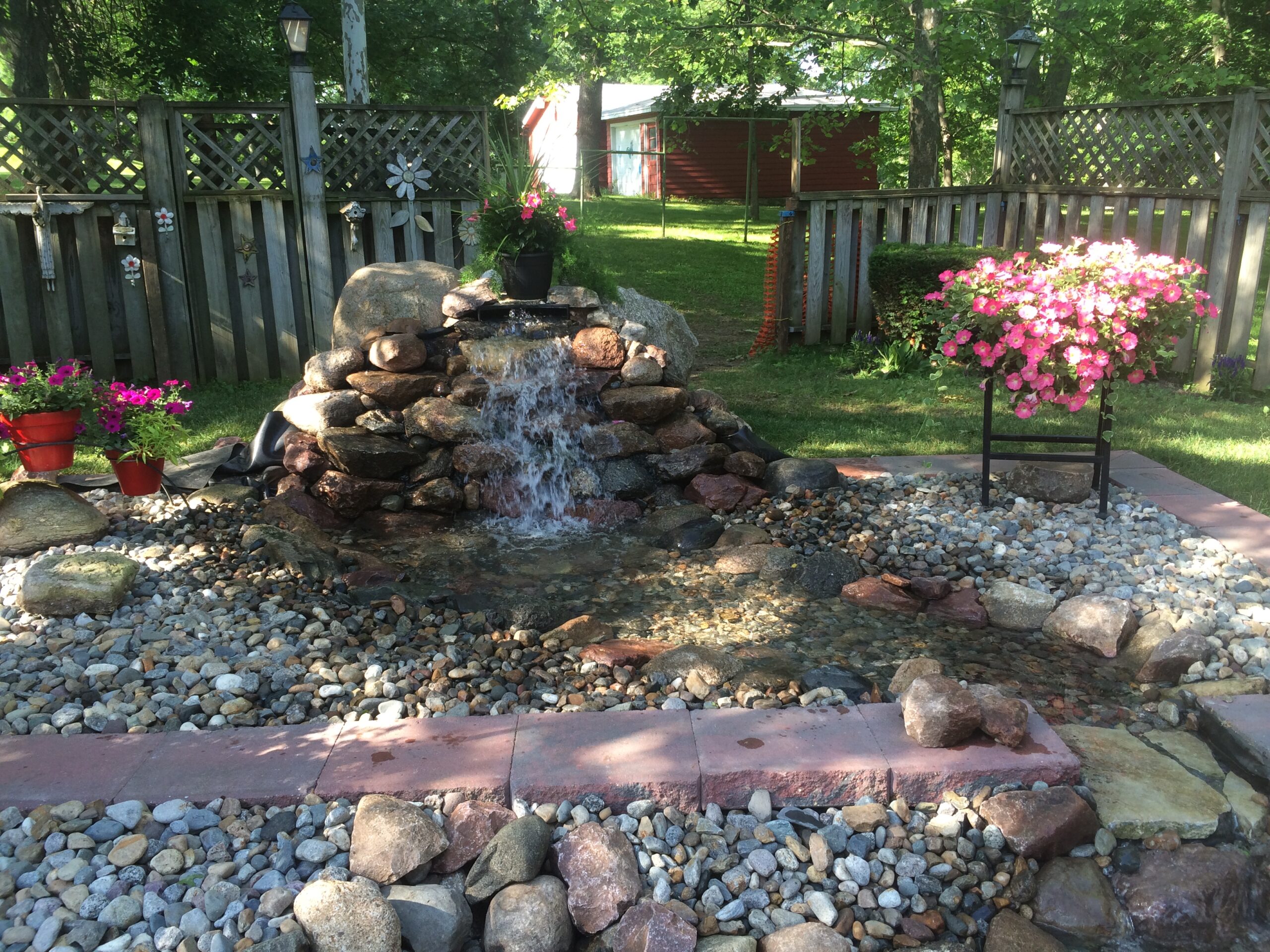 Backyard water feature with cascading waterfall, surrounded by decorative rocks, potted flowers, and a landscaped area, enhancing outdoor living space aesthetics.