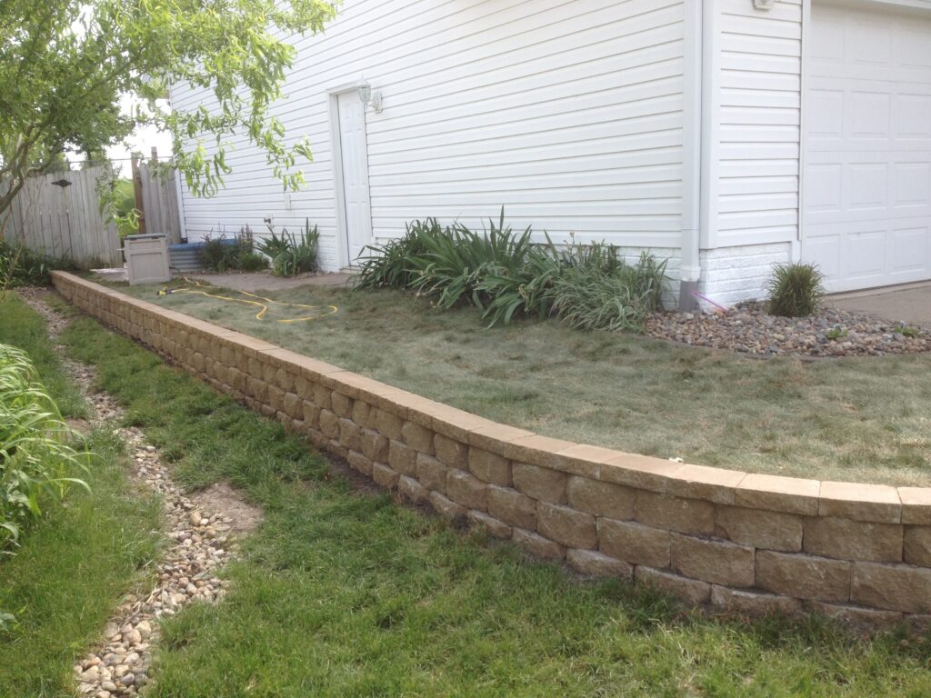 Retaining wall made of stone blocks in a landscaped yard, featuring grass, plants, and a white house exterior in the background, illustrating effective yard design for sloped areas.
