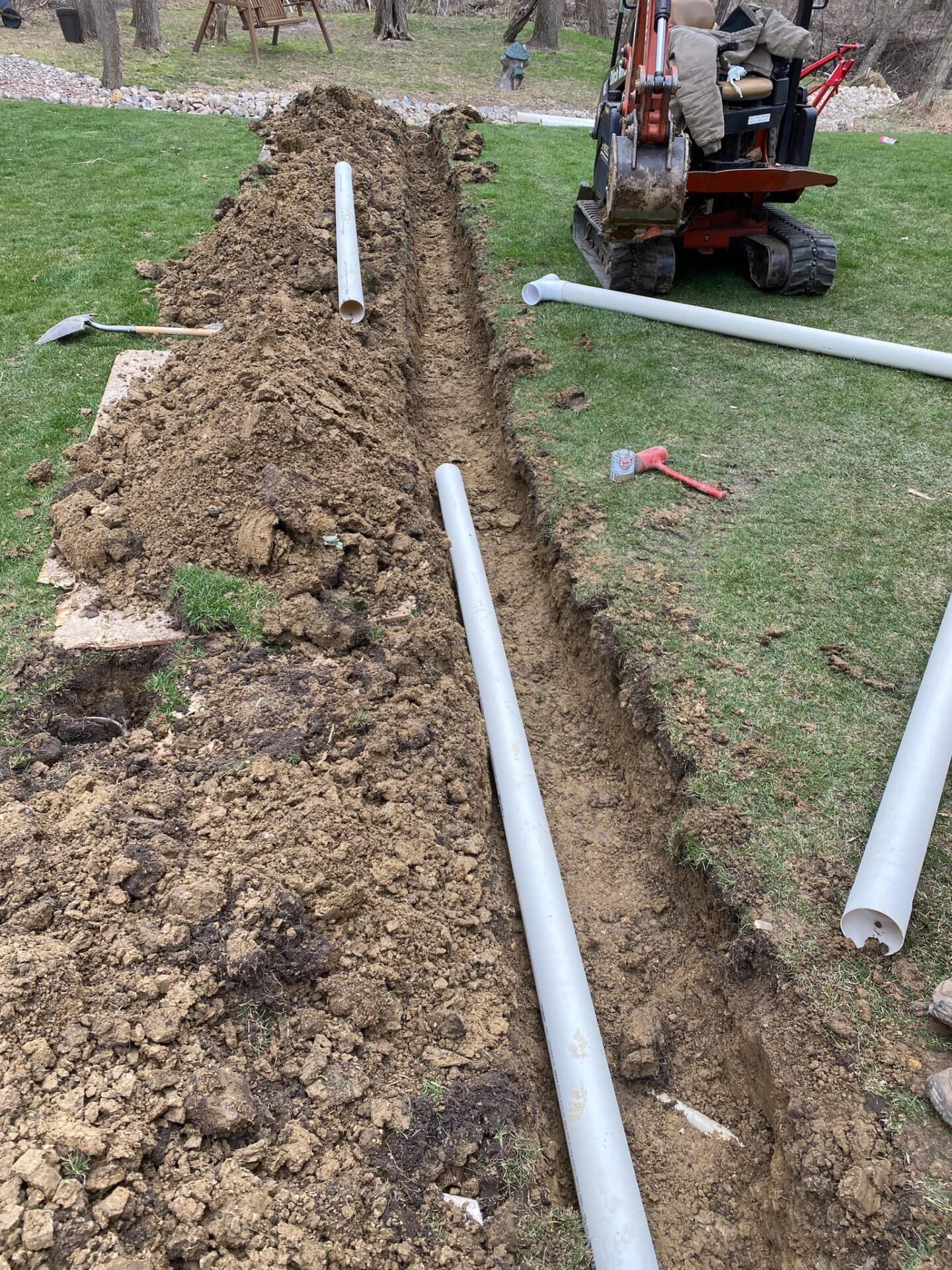 Excavated trench in a yard with gray PVC pipes, landscaping tools, and a mini excavator, illustrating landscaping preparation and installation processes.