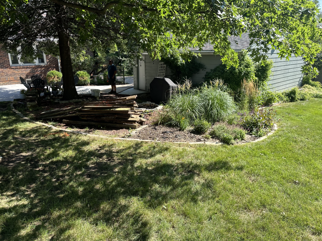 Man standing in a landscaped backyard with natural privacy elements, including a tree and various plants, alongside stacked wooden materials and a patio area.