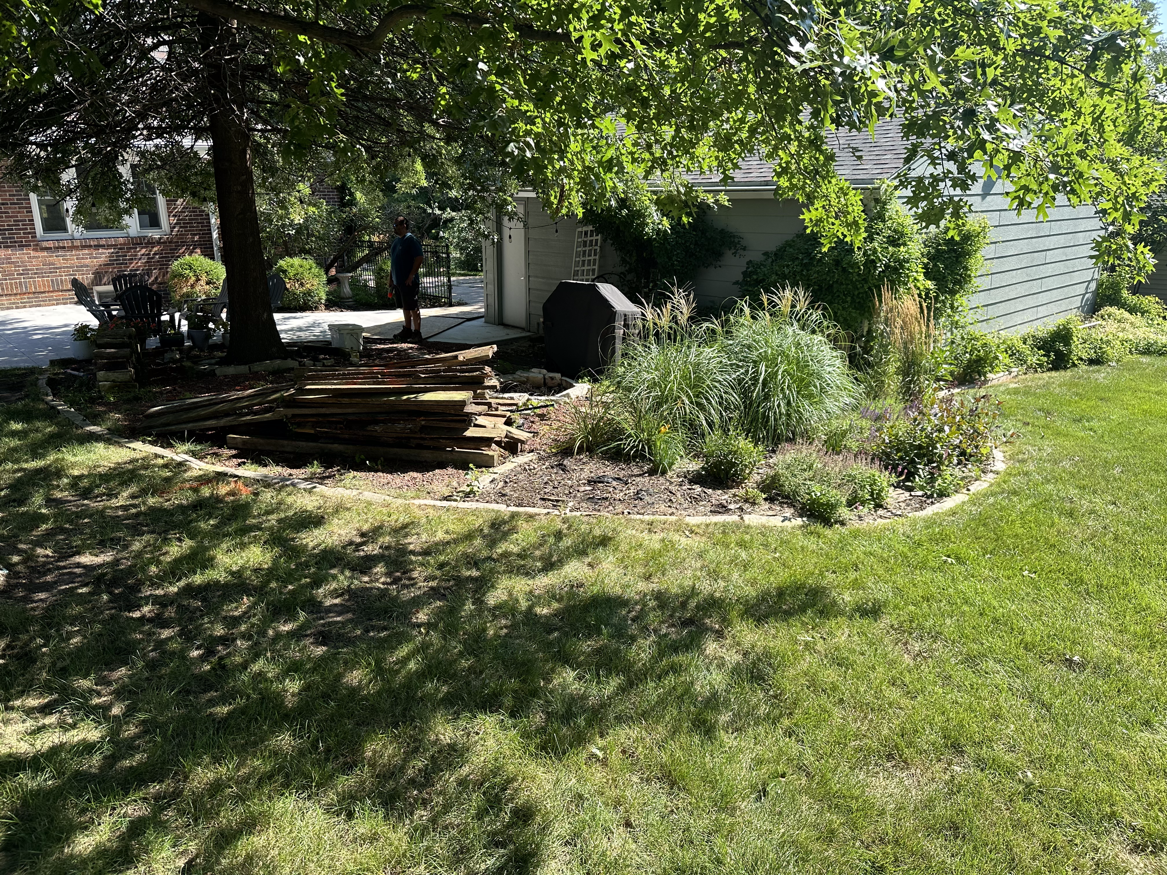 Landscaped outdoor area featuring a man near a stack of wooden planks, surrounded by lush greenery and flower beds, illustrating landscaping work in a residential setting.