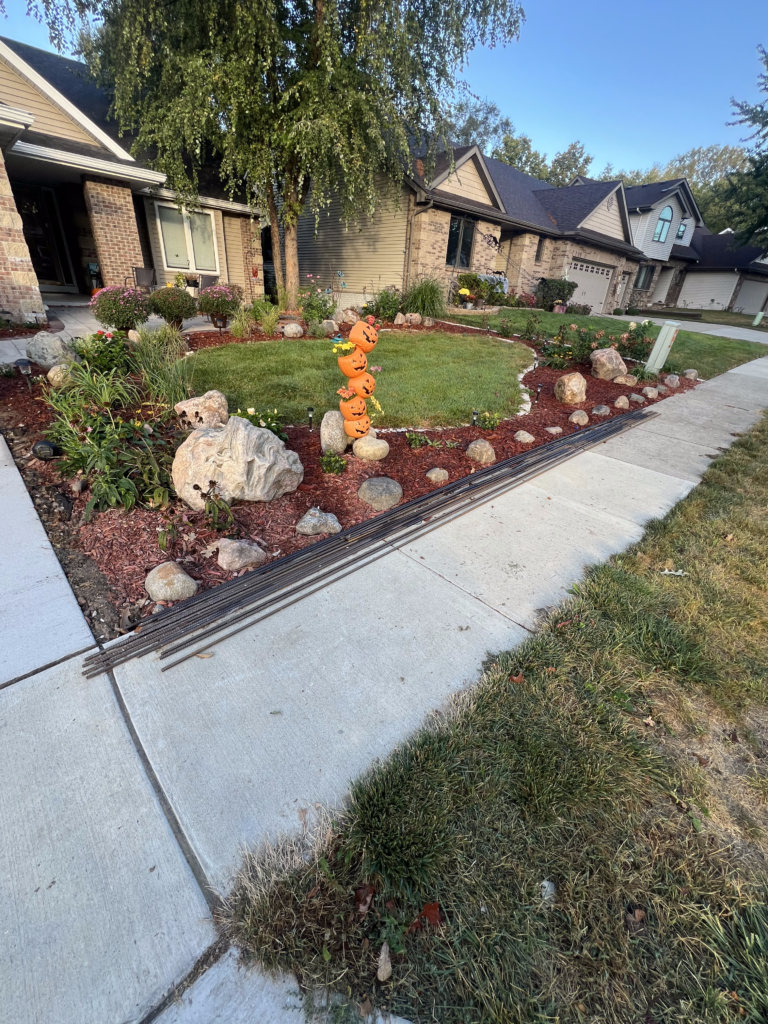 Landscape featuring a well-maintained lawn with decorative pumpkin sculptures, surrounded by colorful flower beds, rocks, and mulch, illustrating seasonal landscaping preparation for fall.