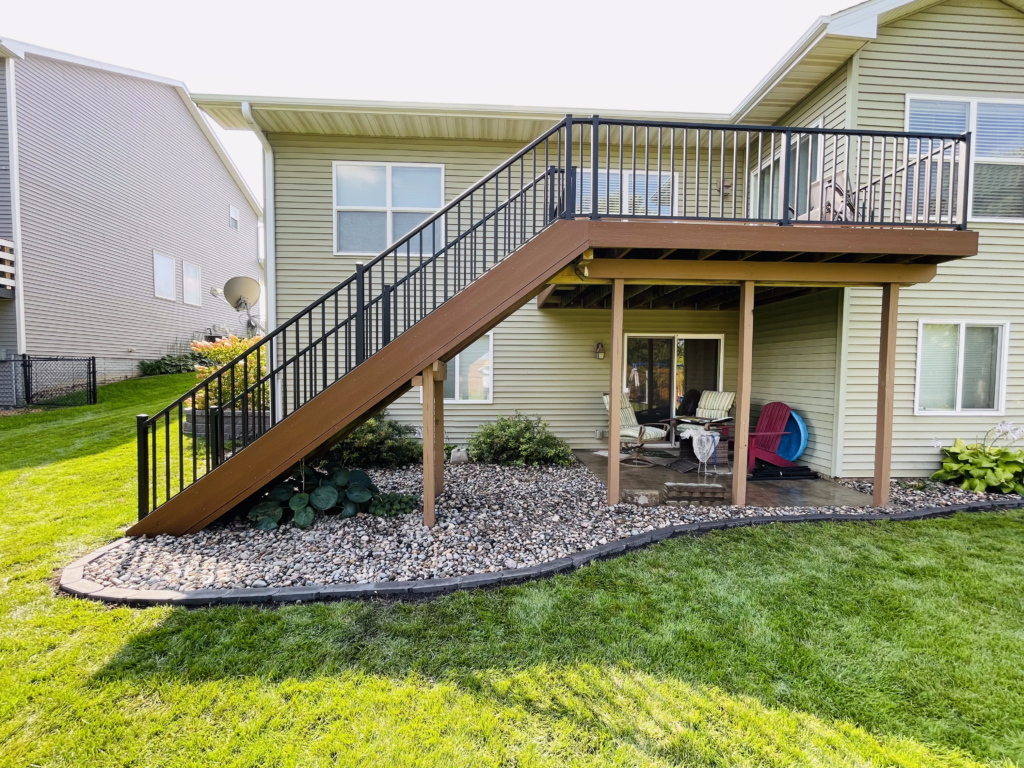 Backyard with a wooden deck and stairs leading to a patio area, featuring landscaped stones and greenery, illustrating a potential space for outdoor gatherings and relaxation, relevant to backyard makeover planning.