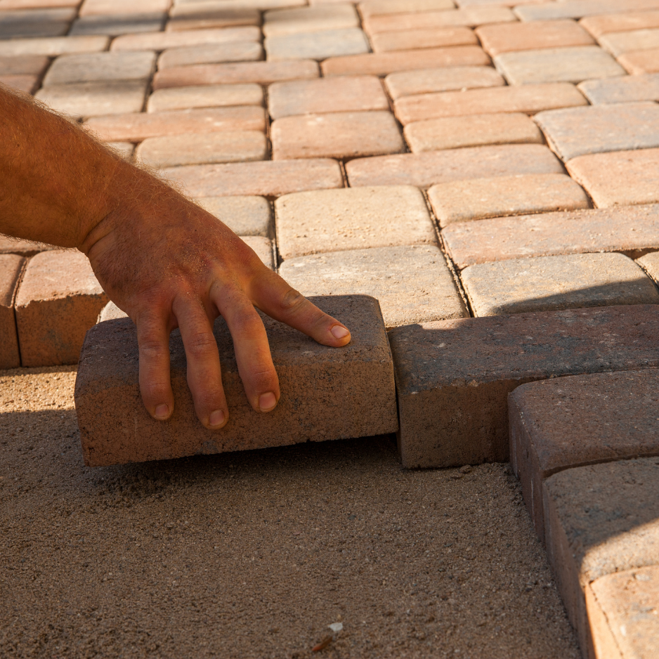 Hand placing a paver brick on a sandy surface, illustrating hardscaping techniques for patio construction and material selection.