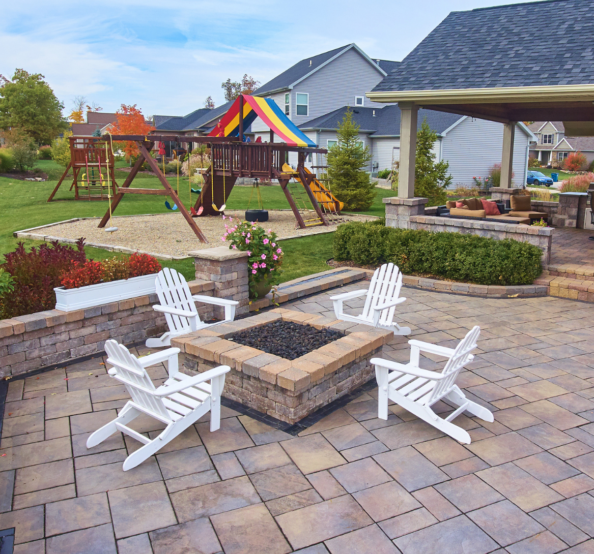 Patio area featuring white Adirondack chairs surrounding a stone fire pit, with a playground set in the background, showcasing a well-designed outdoor space ideal for family activities and landscaping aesthetics.