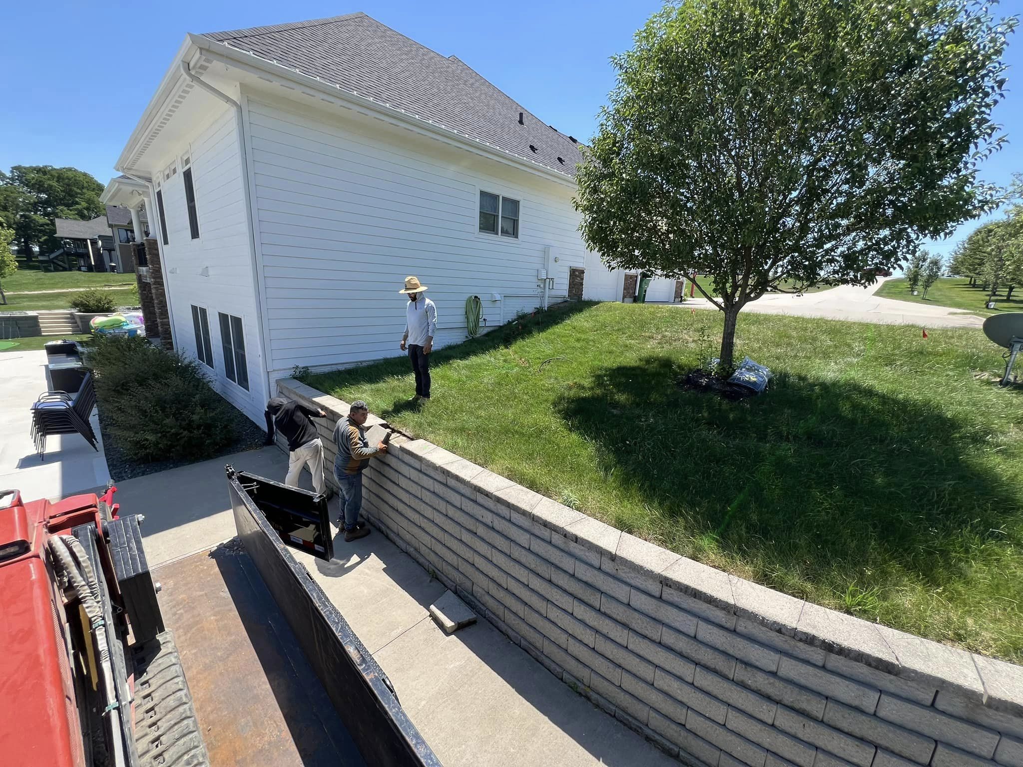 Workers installing a concrete retaining wall in a residential yard, with a house and tree in the background, highlighting hardscaping efforts in landscaping.