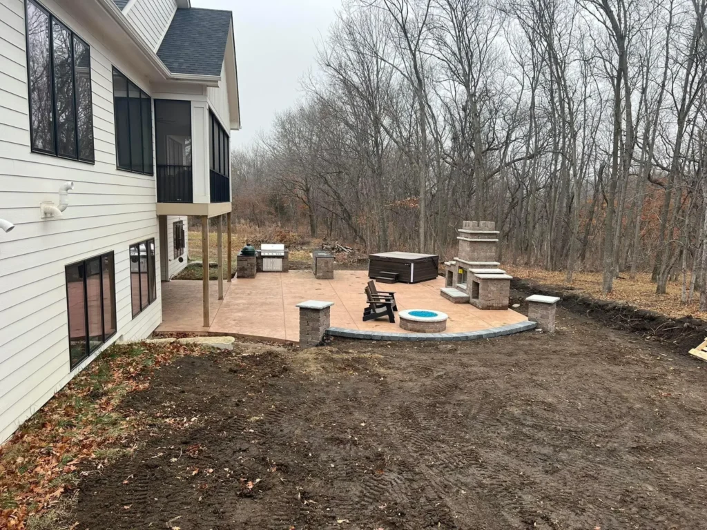 Outdoor living space featuring a patio with a hot tub, seating area, and fireplace, surrounded by bare trees, illustrating low-maintenance landscaping ideal for entertaining in Des Moines.