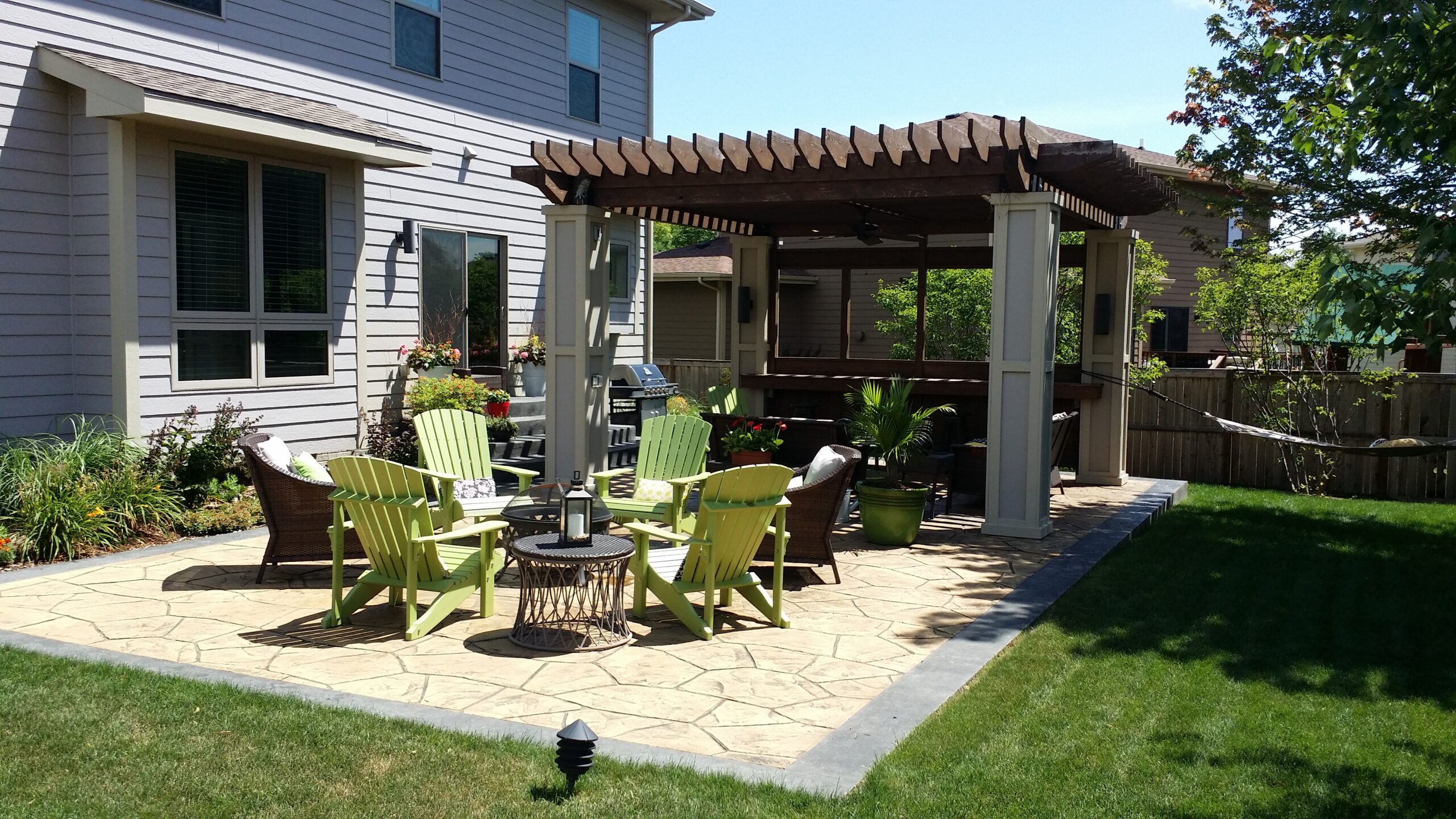 Patio area with green Adirondack chairs, a stone table, and a pergola, surrounded by landscaped garden and home exterior, showcasing outdoor design ideas for family-friendly spaces.